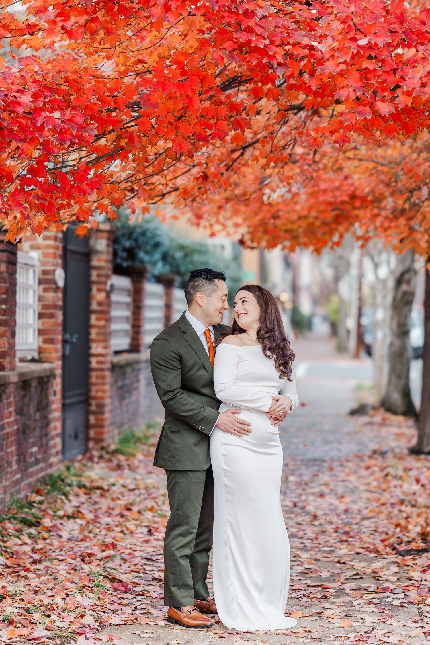 A couple embraces under vibrant red autumn maple trees along a brick wall lined sidewalk with fallen leaves.
