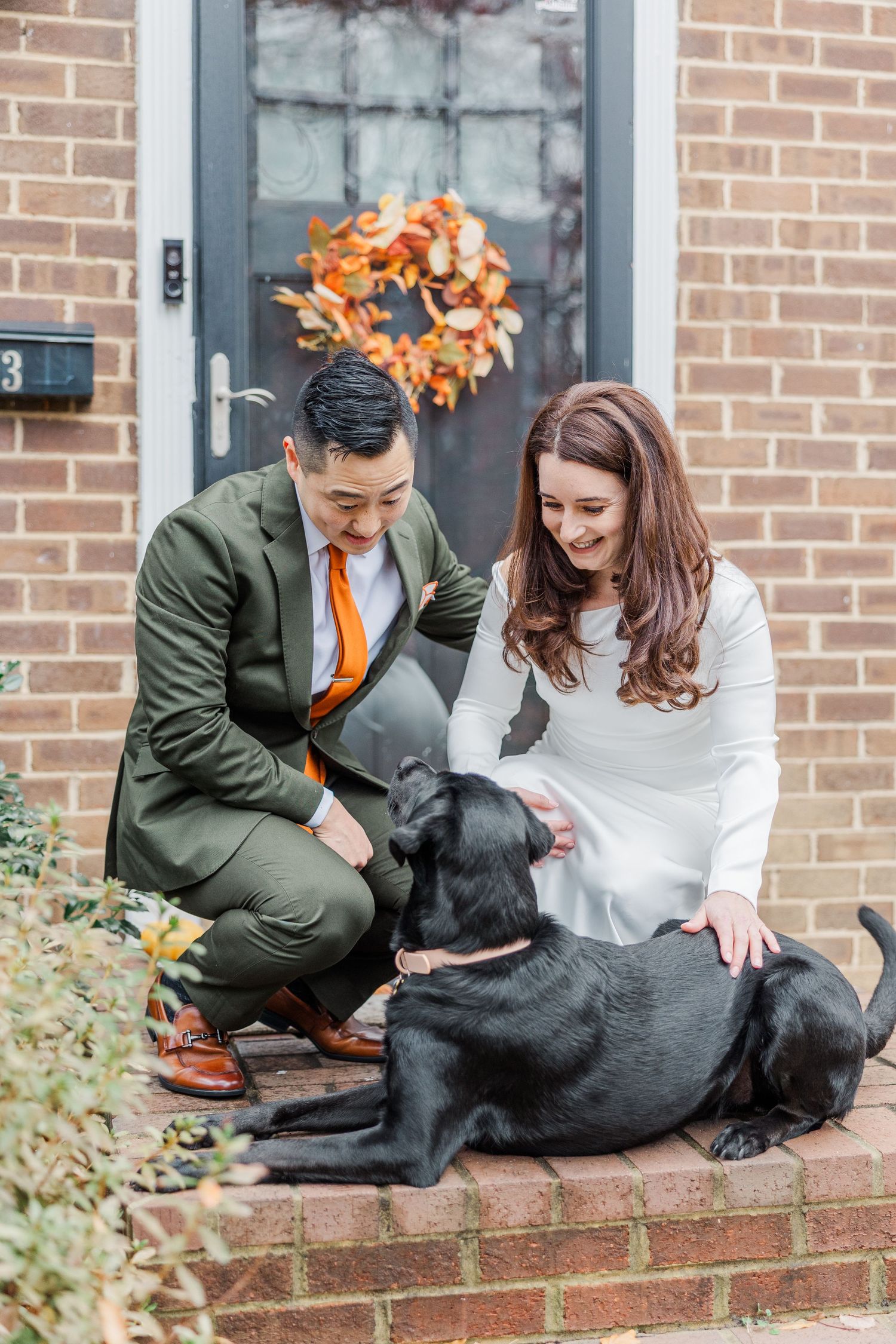 A couple in olive and white attire sits on brick steps with their black dog during an outdoor photoshoot.