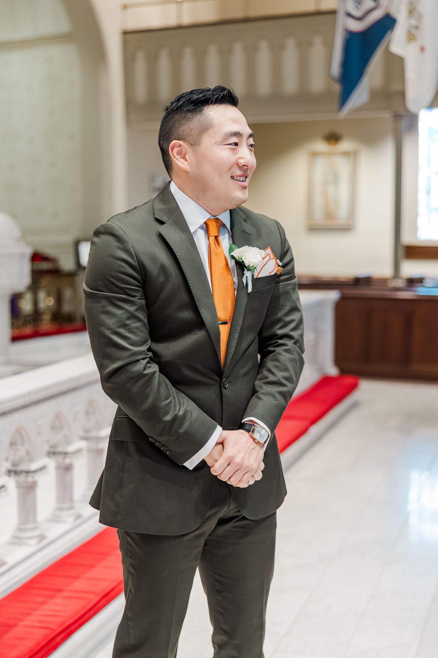 A groomsman in a dark suit and orange tie smiles while standing in a church aisle.
