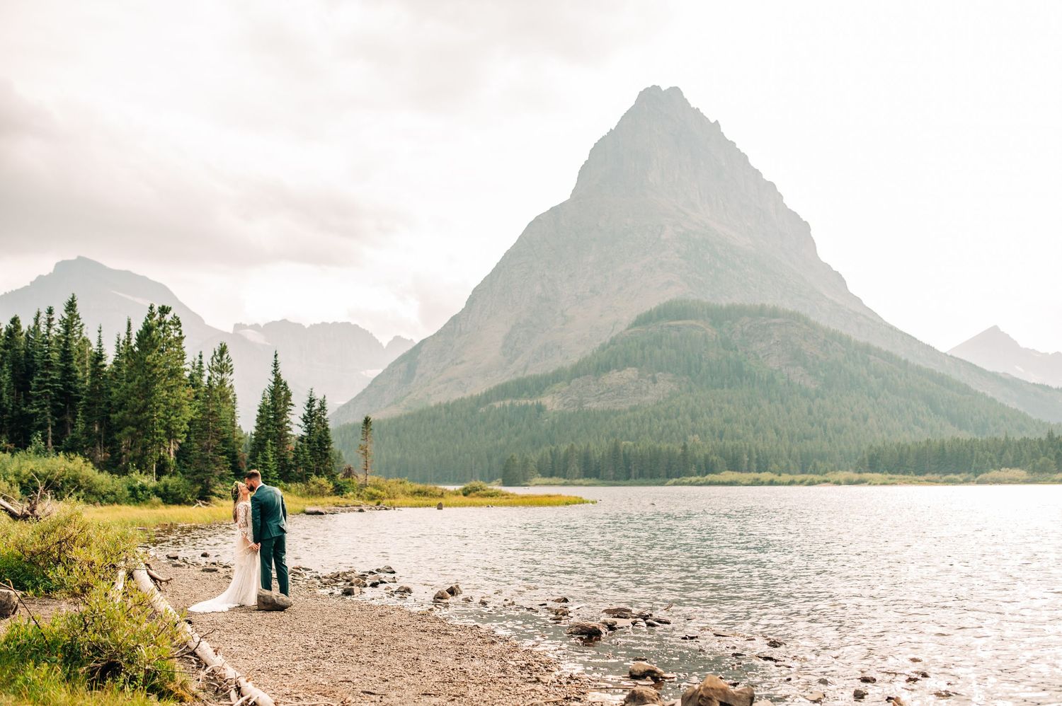A panoramic view of a tranquil mountain lake surrounded by evergreen forest and rocky shoreline at sunset.