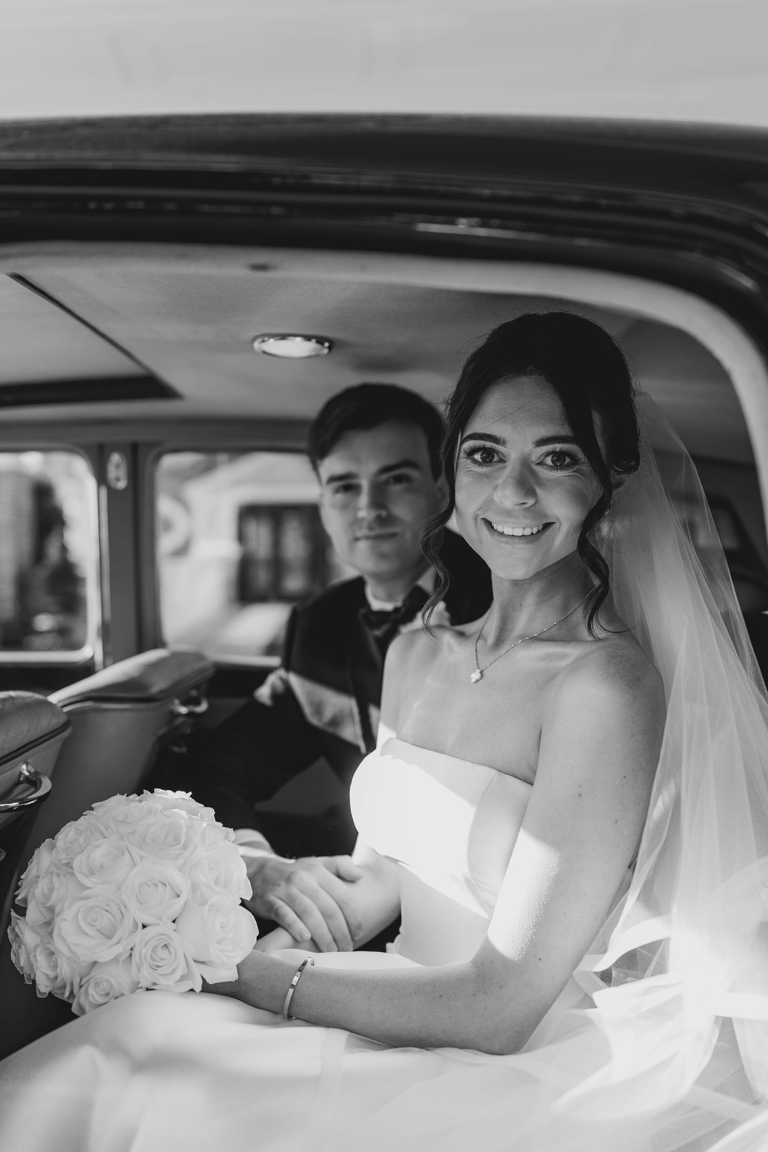 Classic black and white wedding photo of a smiling bride in a white gown holding roses in vintage car interior at the Rockleigh, NJ