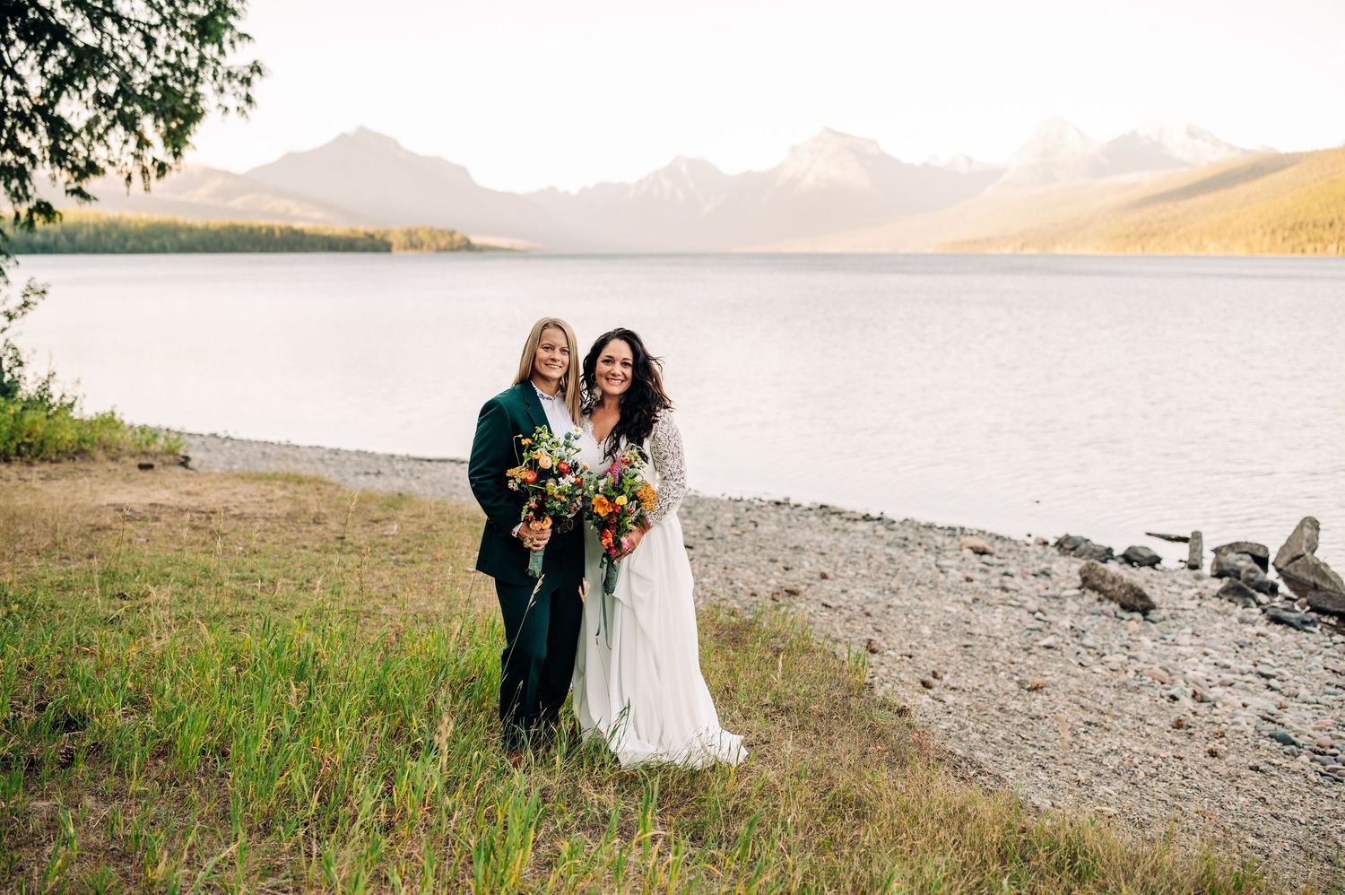 A couple shares a romantic moment on a scenic mountain beach with mountains and water in the background.