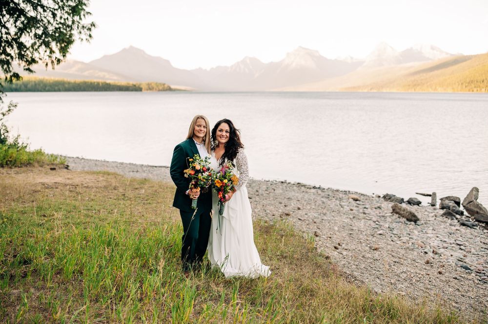 A couple shares a romantic moment on a scenic mountain beach with mountains and water in the background.