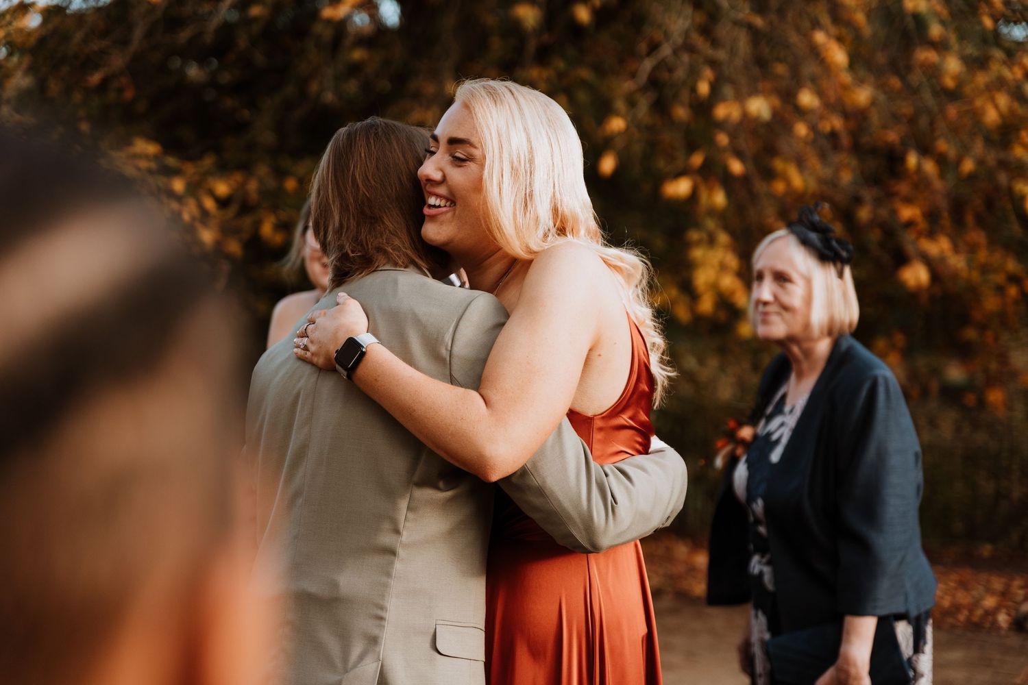 Two friends embrace surrounded by autumn foliage during an outdoor celebration.