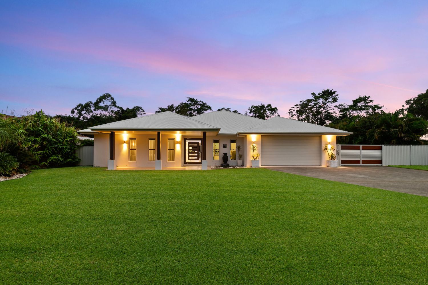 Spacious single-story home with wide green lawn, warm interior lighting, and lush trees at dusk.