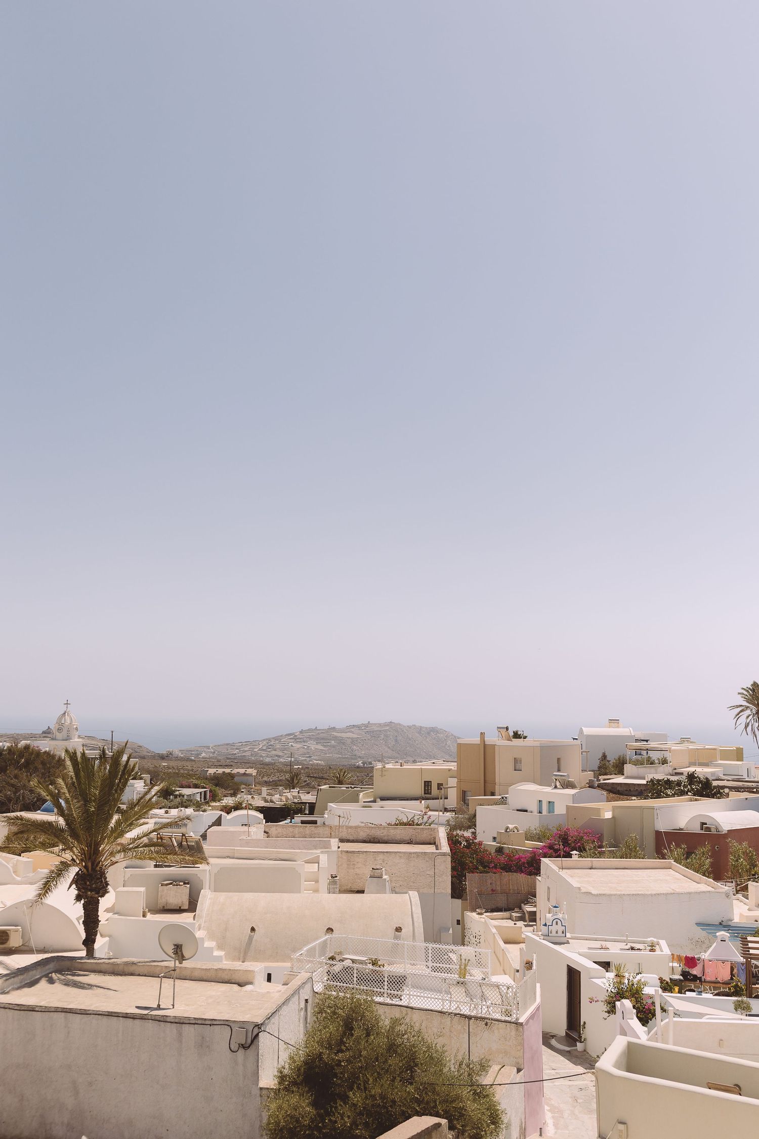 Panoramic view of white Mediterranean buildings with flat roofs and palm trees against a cloudy sky in Santorini.