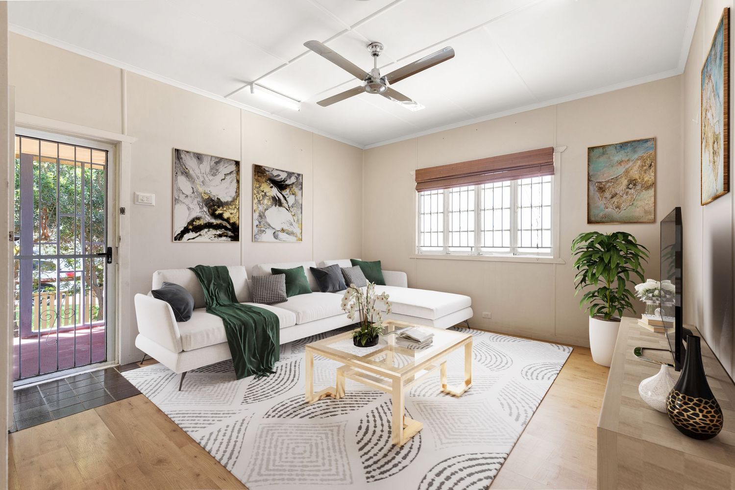 Bright living room with white sofa, ceiling fan, hardwood floors, and large windows with garden views.