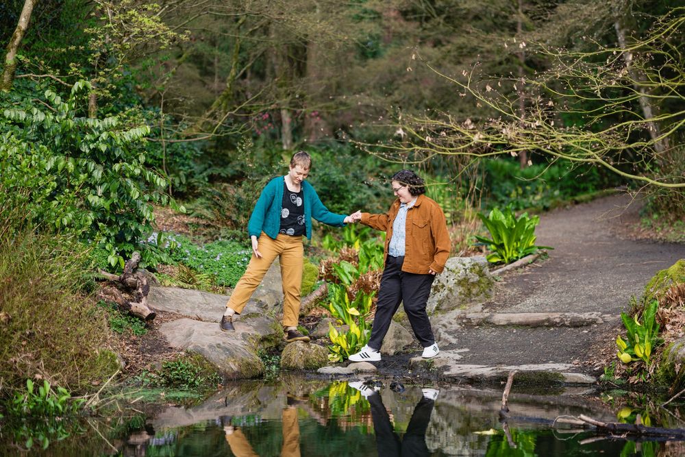 Two women help each other across stepping stones over a reflective pond in a lush green garden.