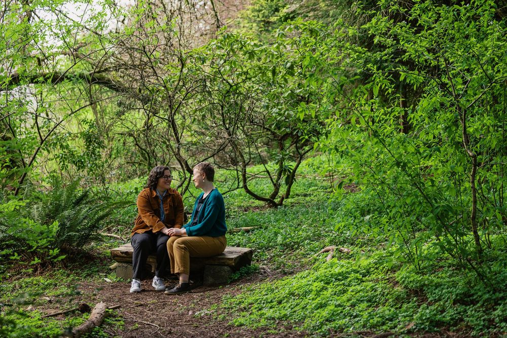 Two people sitting on a log bench sharing an intimate conversation in a lush green woodland setting.