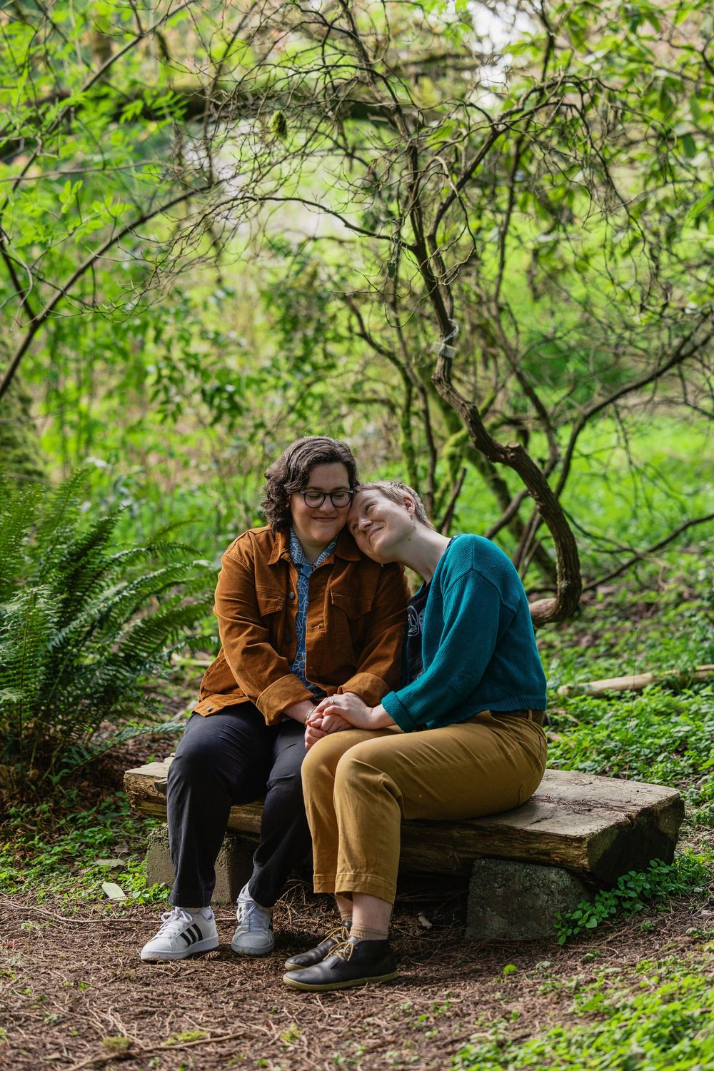Two women sitting on a stone bench in a garden, one resting her head on the other's shoulder tenderly.