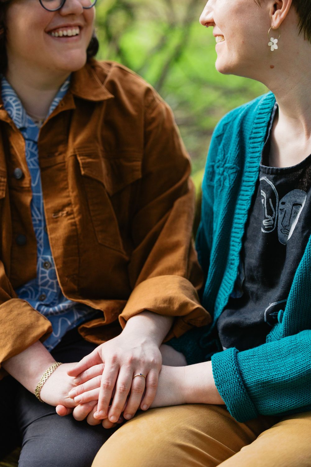 Close-up of two women holding hands while sitting together, one wearing a brown jacket and the other teal.