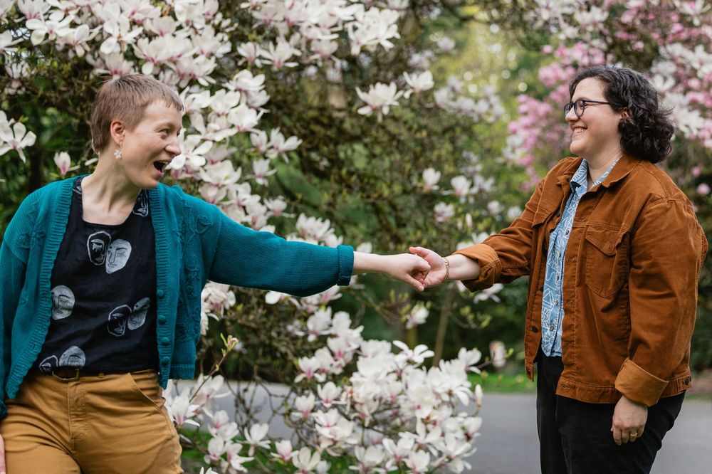 Two women laugh and hold hands in front of flowering magnolia blossoms.