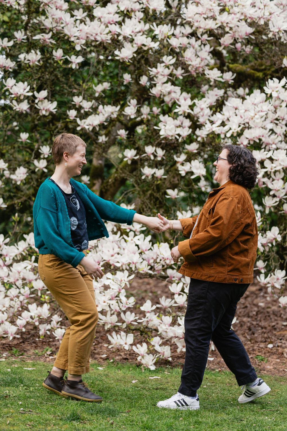 Two women dance and hold hands on green grass beside a magnolia tree.