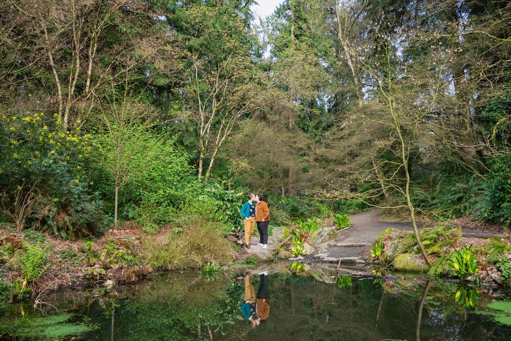 Couple standing on stone bridge reflected in tranquil pond surrounded by lush green trees in a botanical garden.