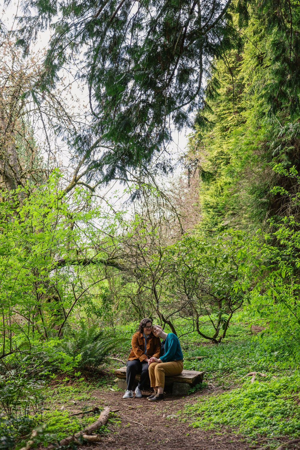 Couple sitting close together on a rustic stone bench surrounded by vibrant green forest trees.