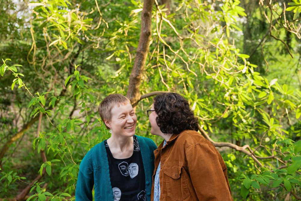 Two women smiling and gazing at each other surrounded by bright green spring foliage in a forest.