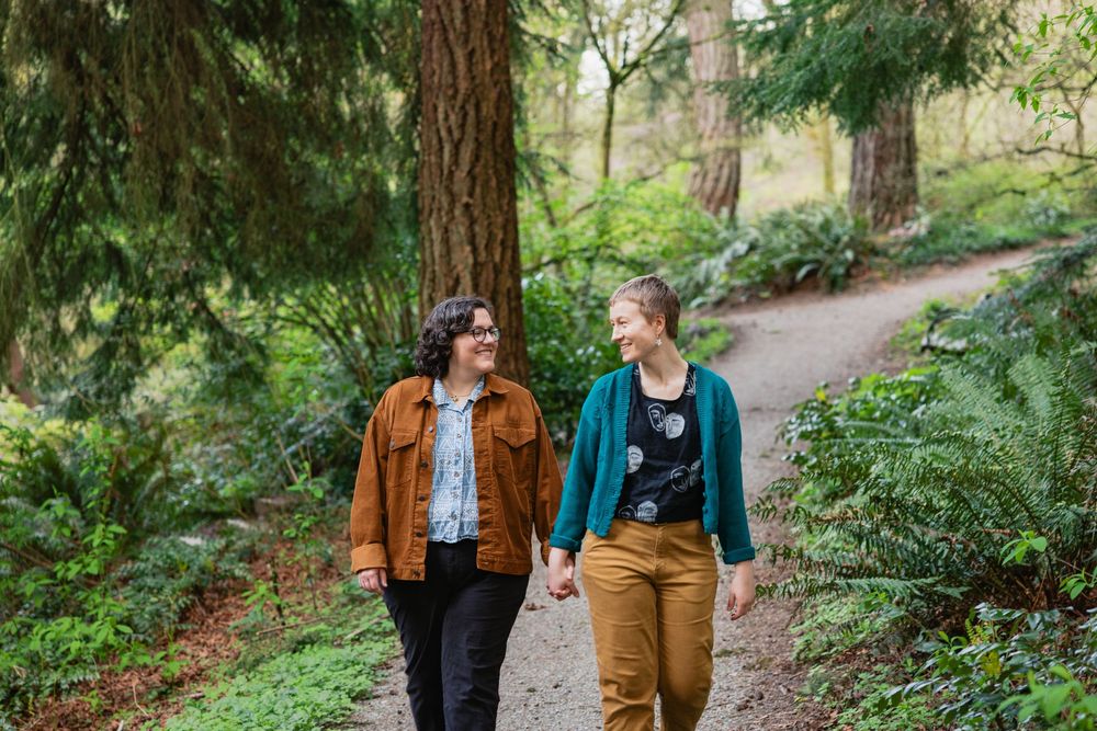 Two women walking hand in hand along a lush green garden path lined with tall trees.