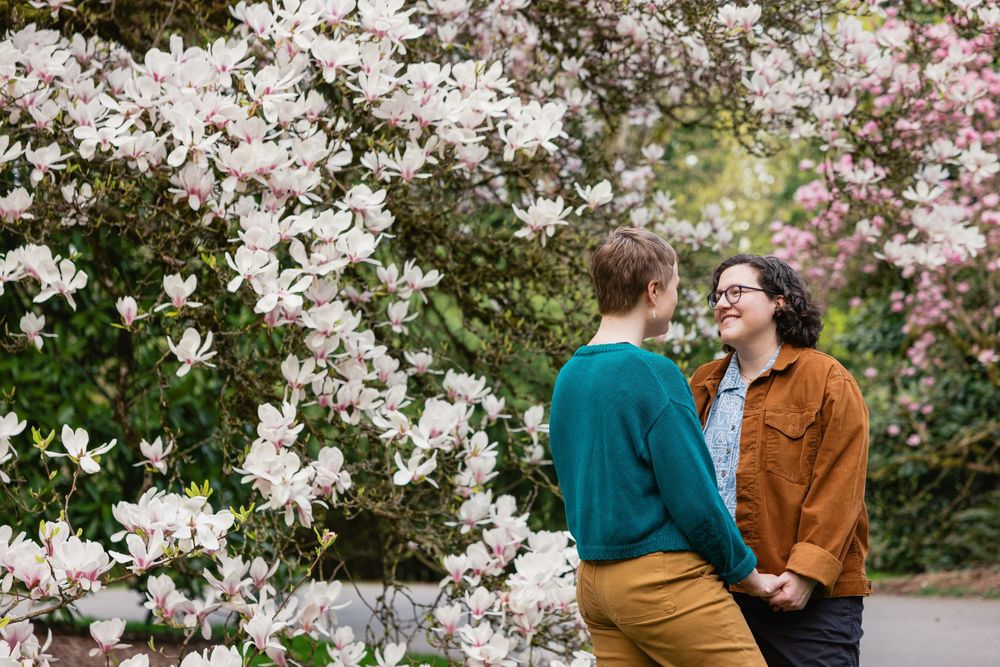 Two women holding hands and smiling at each other beside a large blooming magnolia tree in spring.