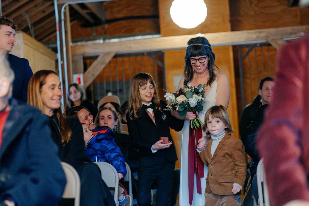 Bride in white dress with bouquet walking down aisle with children in rustic barn ceremony.