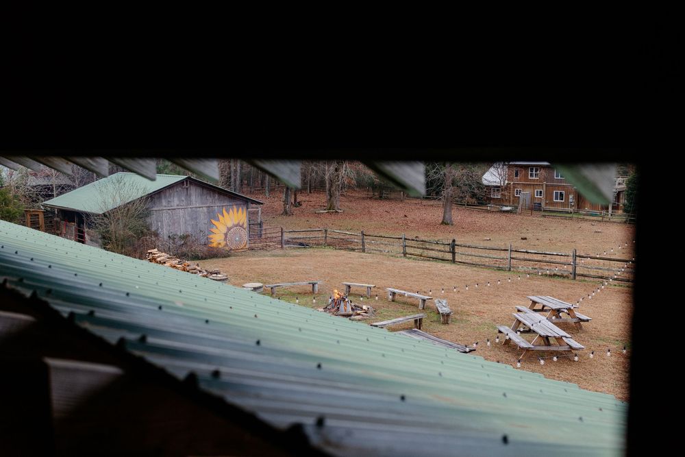 Aerial view through gap of rustic farm with wooden barns, fenced corral, and picnic tables below.