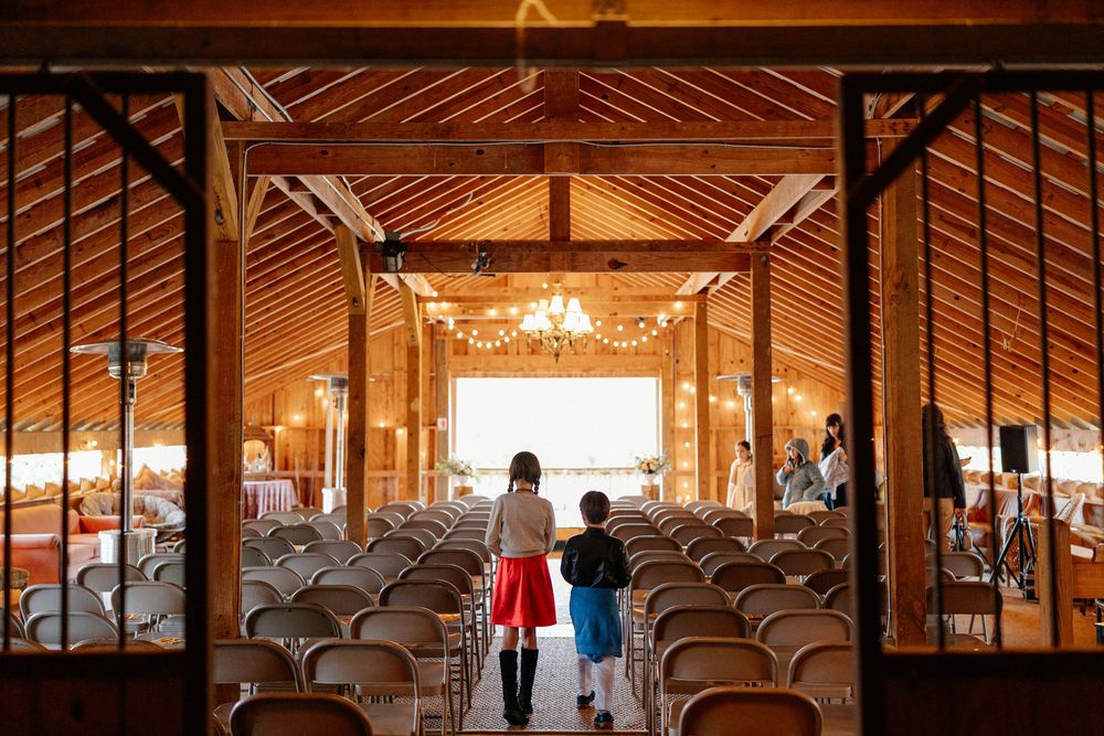 Two children walking hand in hand into rustic barn wedding venue with rows of white chairs.