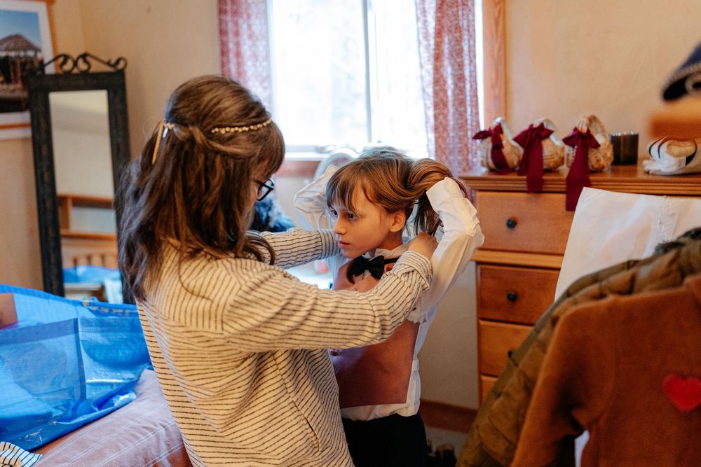 Young girl helping fix hair accessory on smaller child in cozy bedroom during wedding preparation.