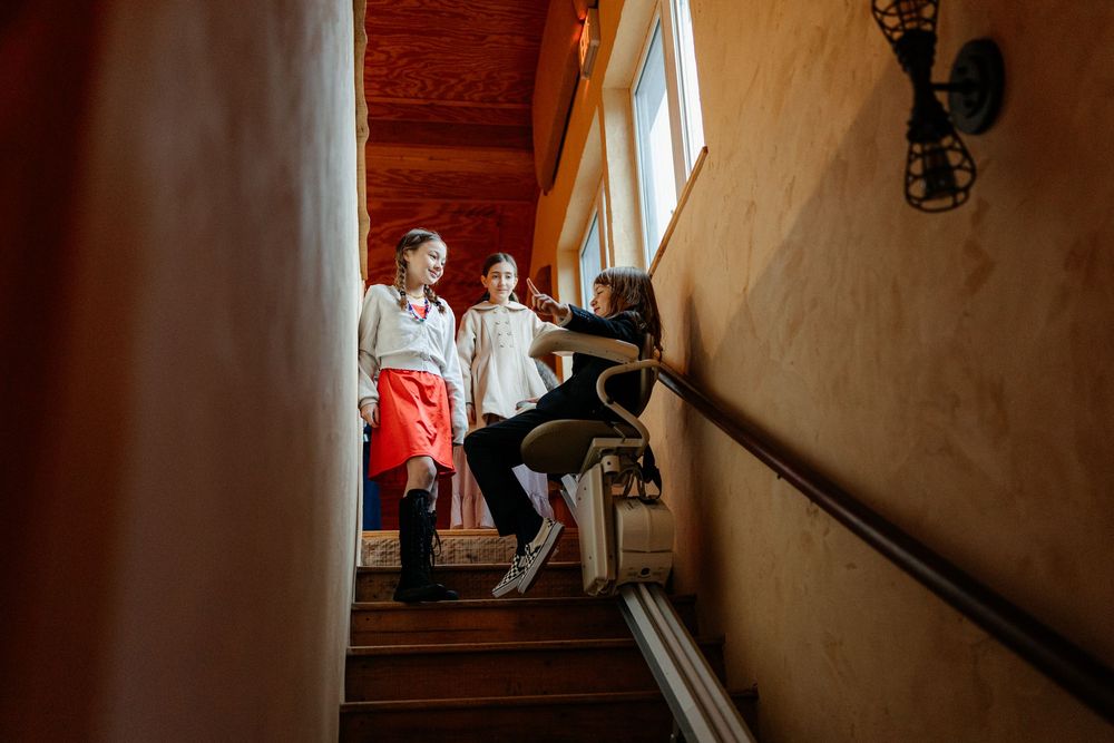 Children watching woman ride stairlift down wooden staircase inside warm rustic home.