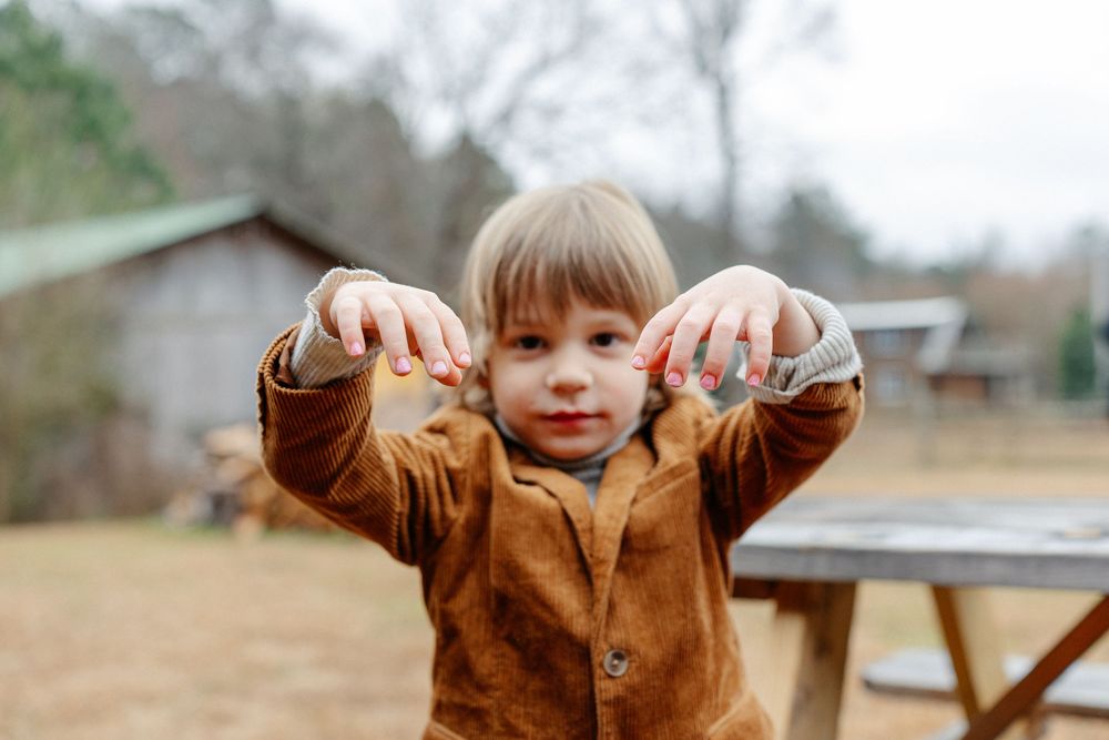 Adorable young child in brown jacket holding out hands outdoors at a rustic wedding venue.