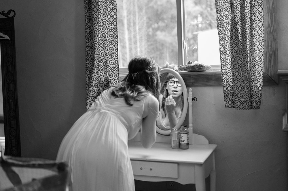 Black and white photo of bride applying makeup at a vintage vanity mirror before wedding.