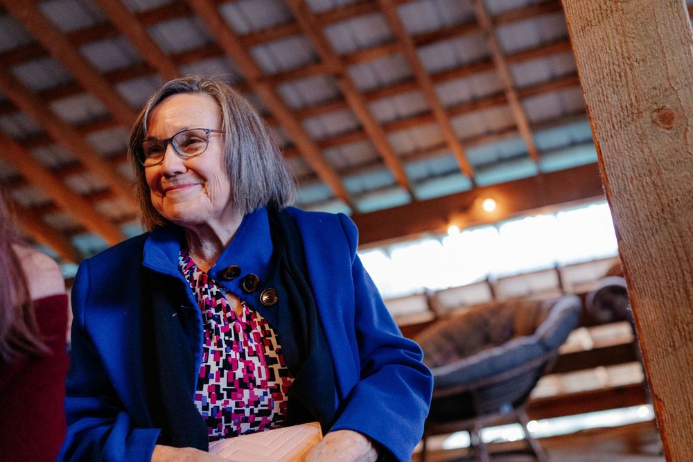 Elegant older woman in blue blazer smiling warmly inside rustic wooden barn wedding venue.