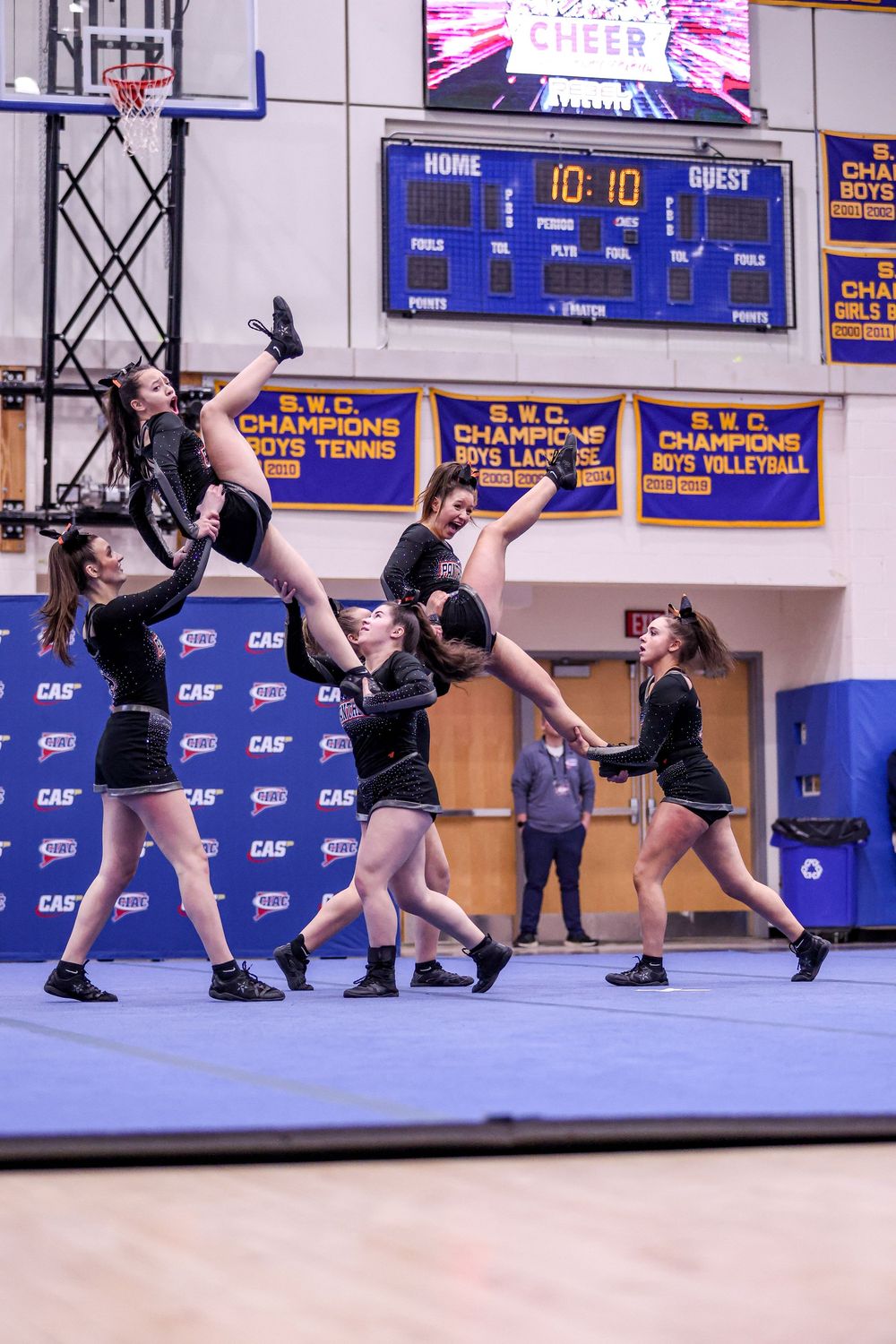 Cheerleaders in black uniforms perform a stunt routine on a blue mat in a school gymnasium during a competition.