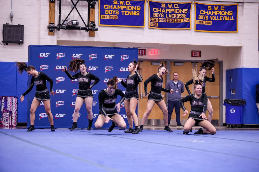 Cheerleading team performing dynamic routine on blue mat in school gymnasium with championship banners on wall.