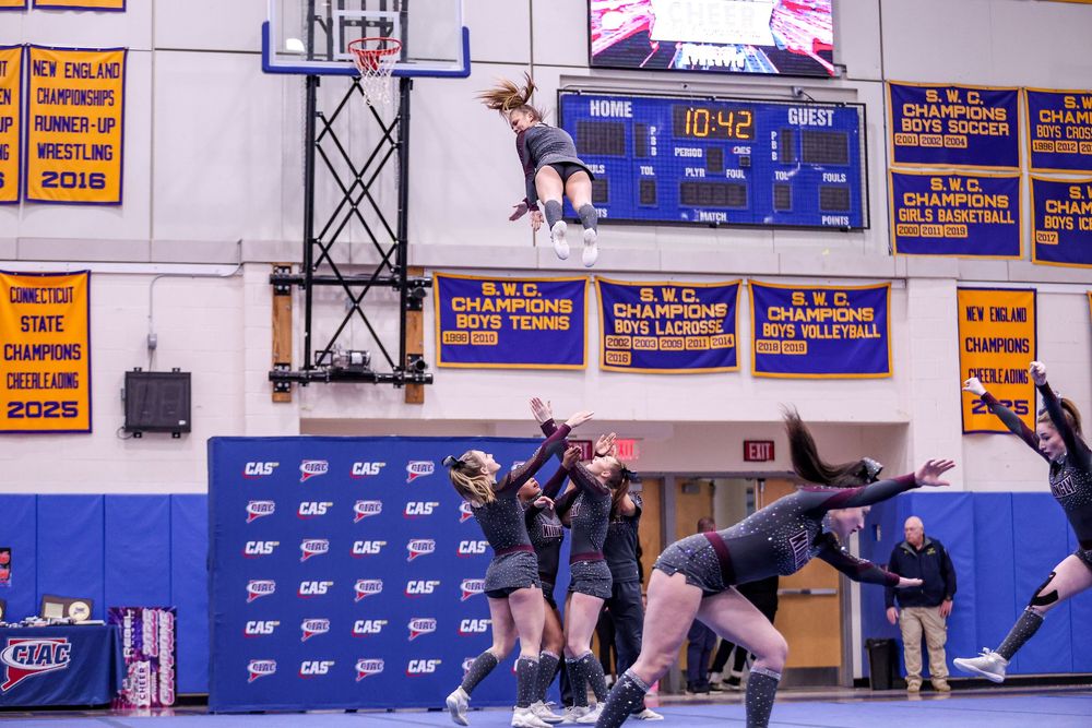 High school cheerleaders performing dynamic stunts in a gymnasium decorated with championship banners during a sports event.