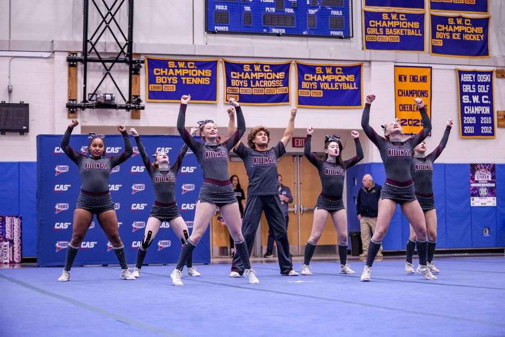 Cheerleading squad performing a jump routine in a gymnasium with championship banners on the wall.