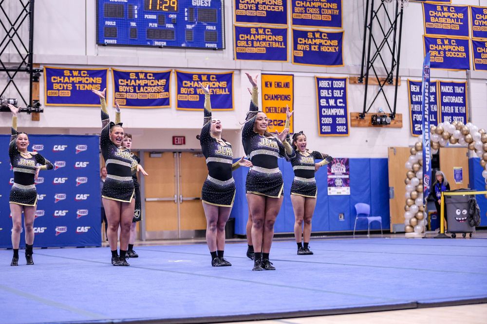 Cheerleaders perform a routine at an indoor competition with blue mats, trophy displays, and championship banners on the wall.