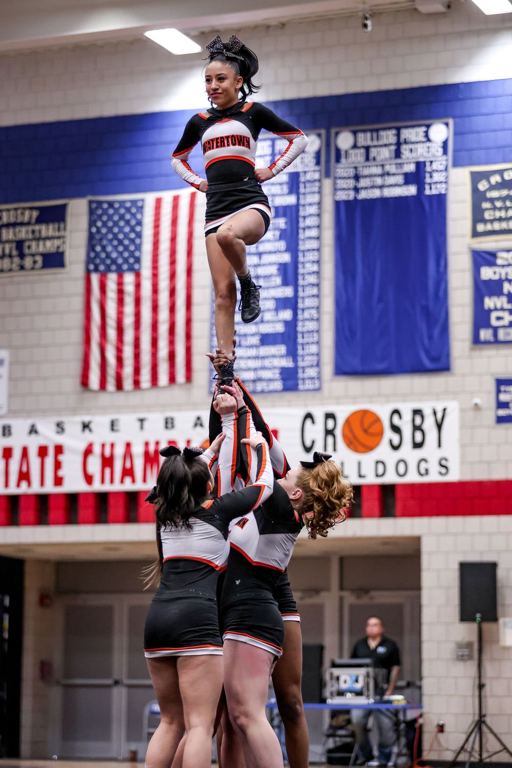 Cheerleaders perform stunts and formations in a school gym with American flags and championship banners on display.