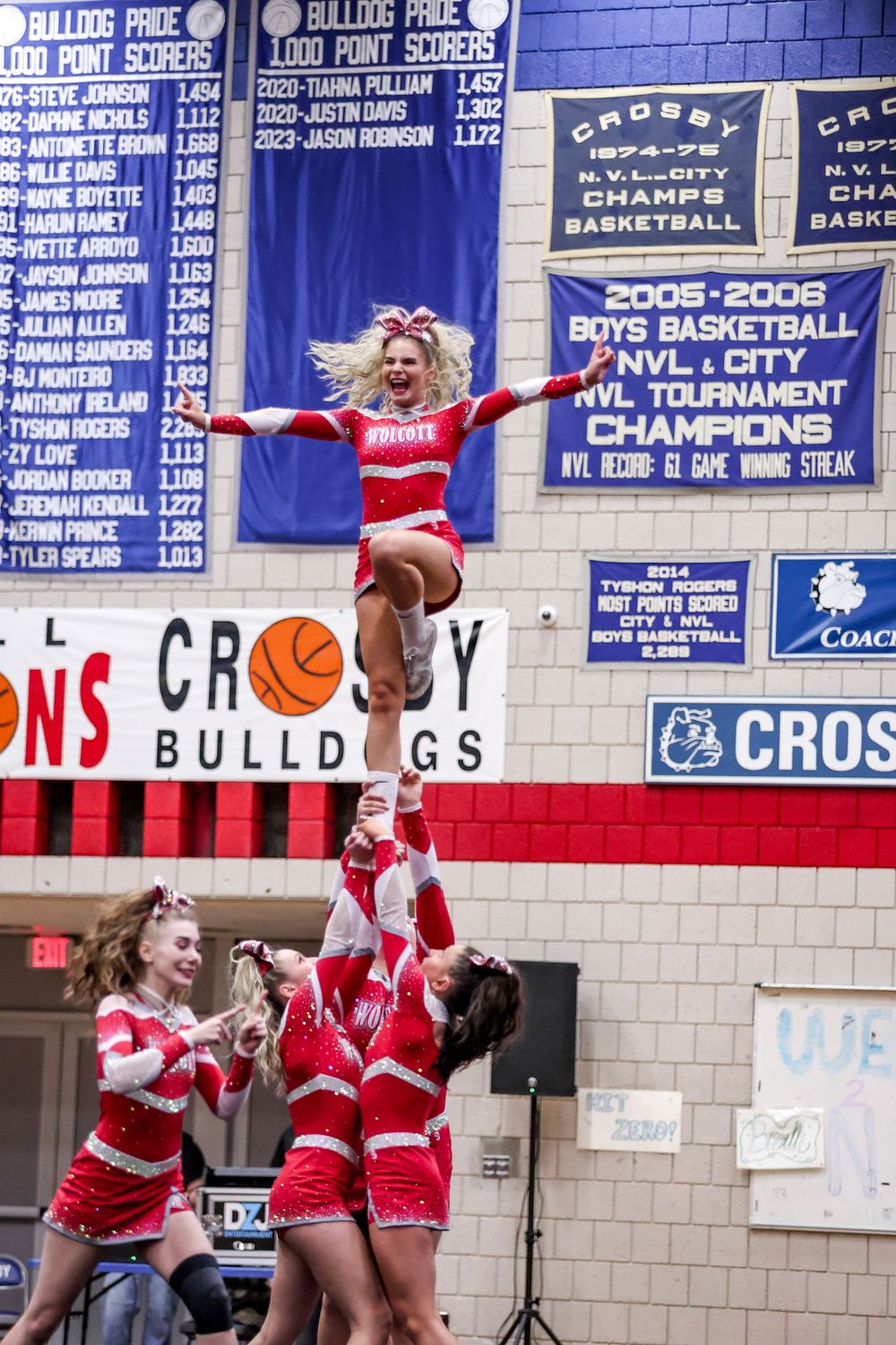 Cheerleaders in red and white uniforms perform synchronized jumps and stunts during a high school basketball game.