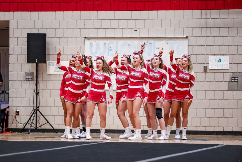 A cheerleading squad in red and white striped uniforms performs a synchronized routine in front of a brick wall.