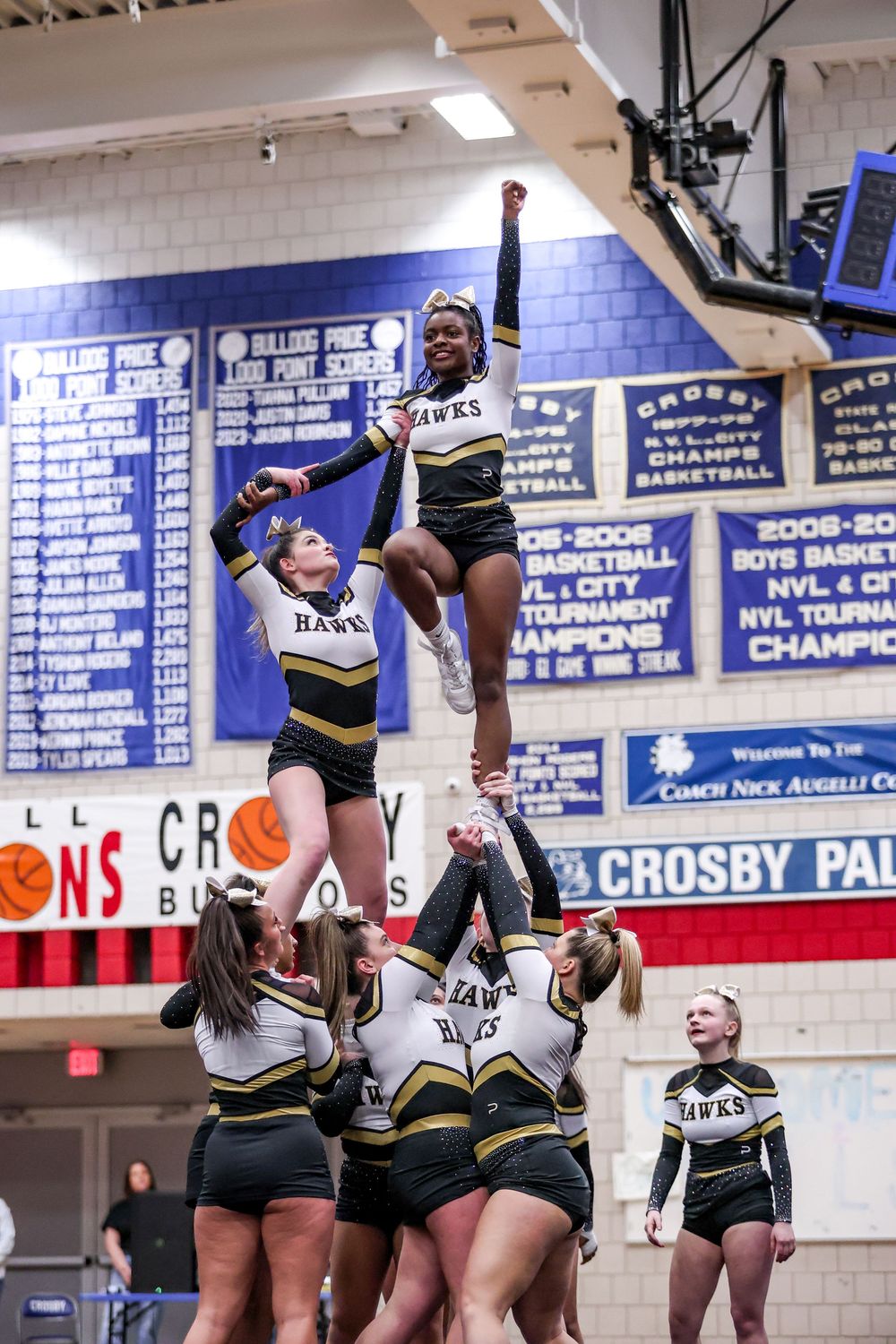 Cheerleaders perform a high-flying stunt in black and white uniforms during a competition at a high school gymnasium.