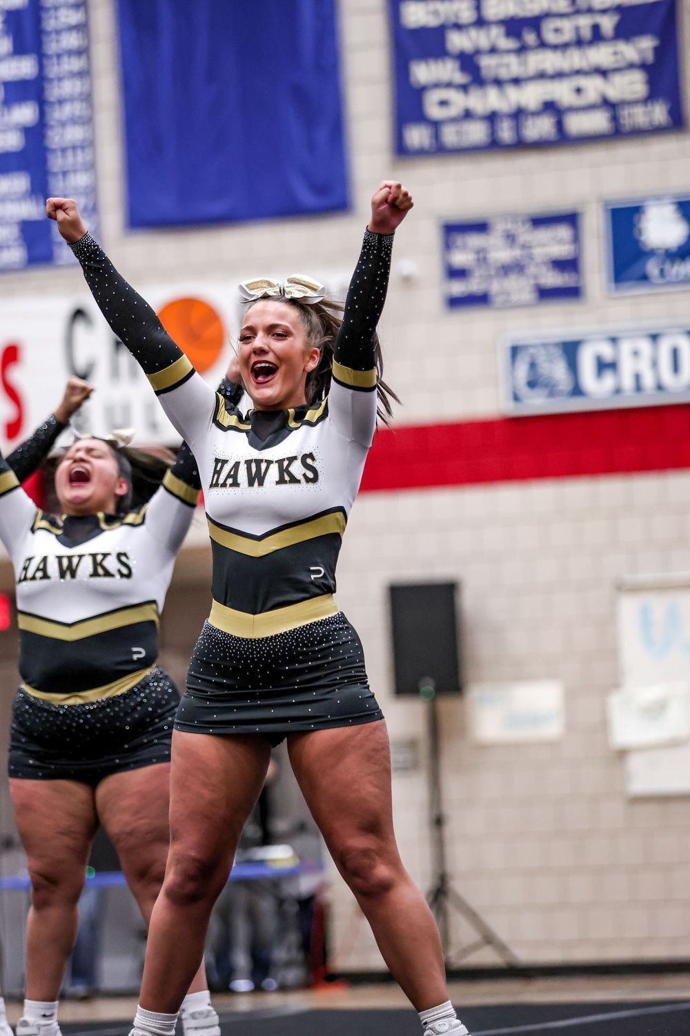 High school cheerleaders in black and gold Hawks uniforms celebrate energetically during a performance.