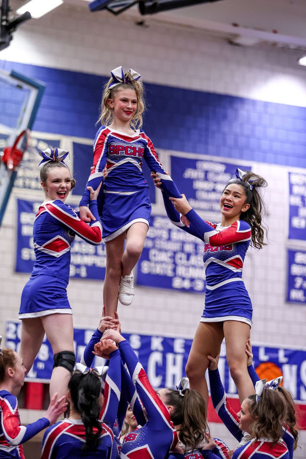 Cheerleaders performing synchronized stunts and lifts during a high school pep rally in a blue-walled gymnasium.
