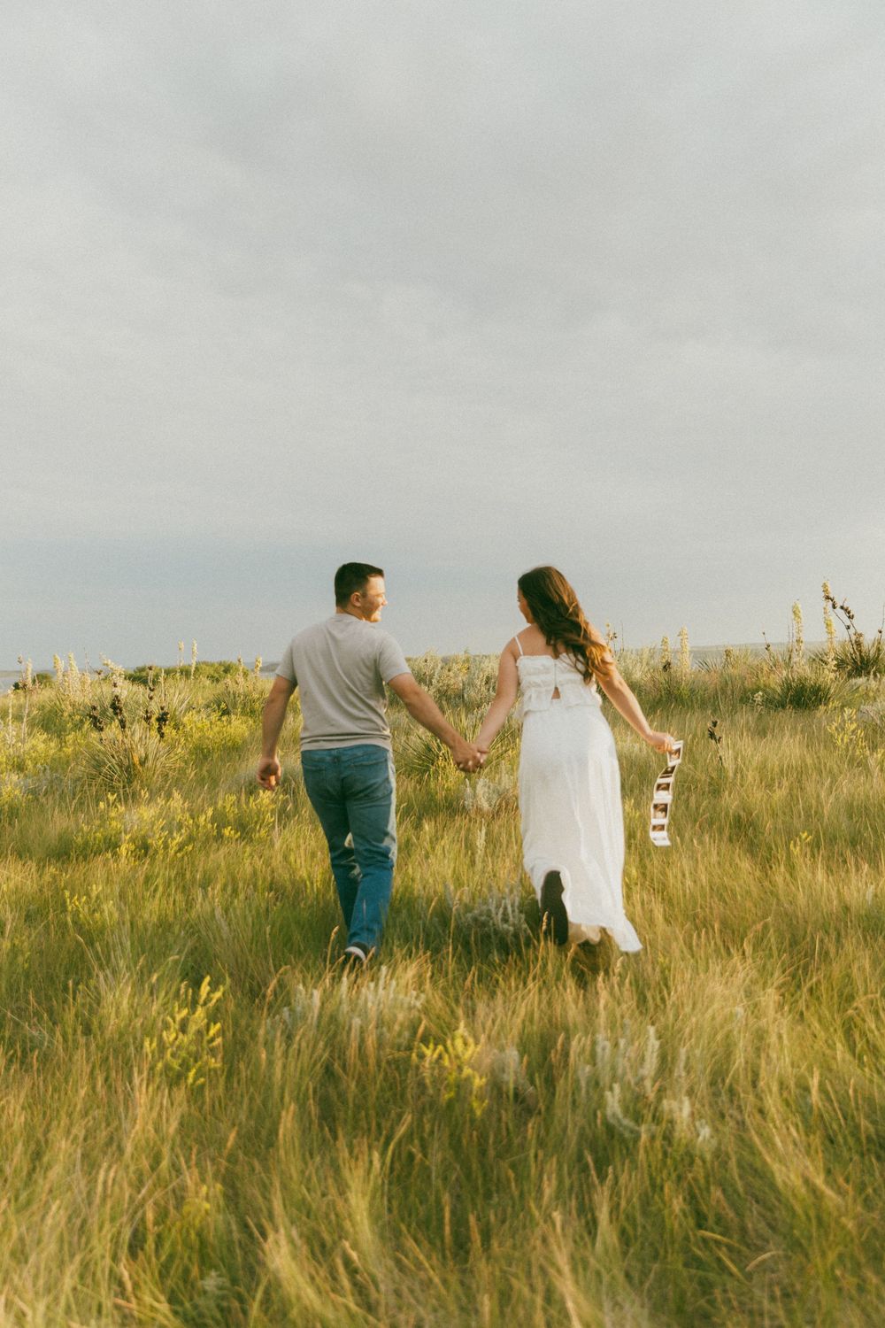A couple walks hand in hand through a golden field of tall grass on a cloudy day.