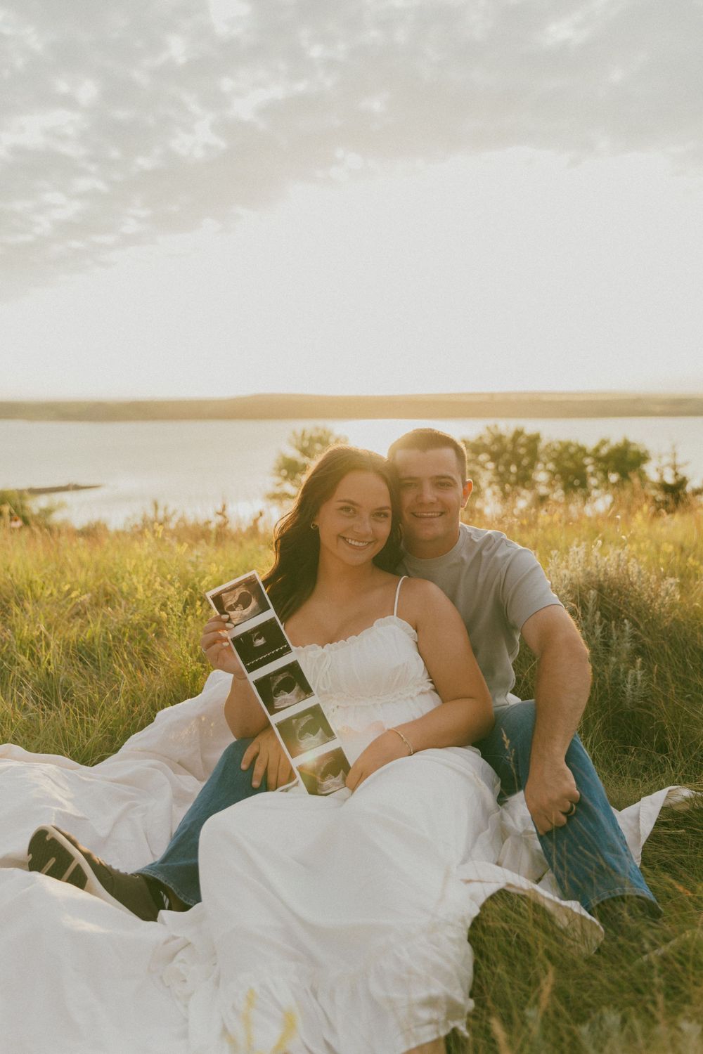 A couple sits in a grassy field at sunset holding ultrasound photos beside a lake.