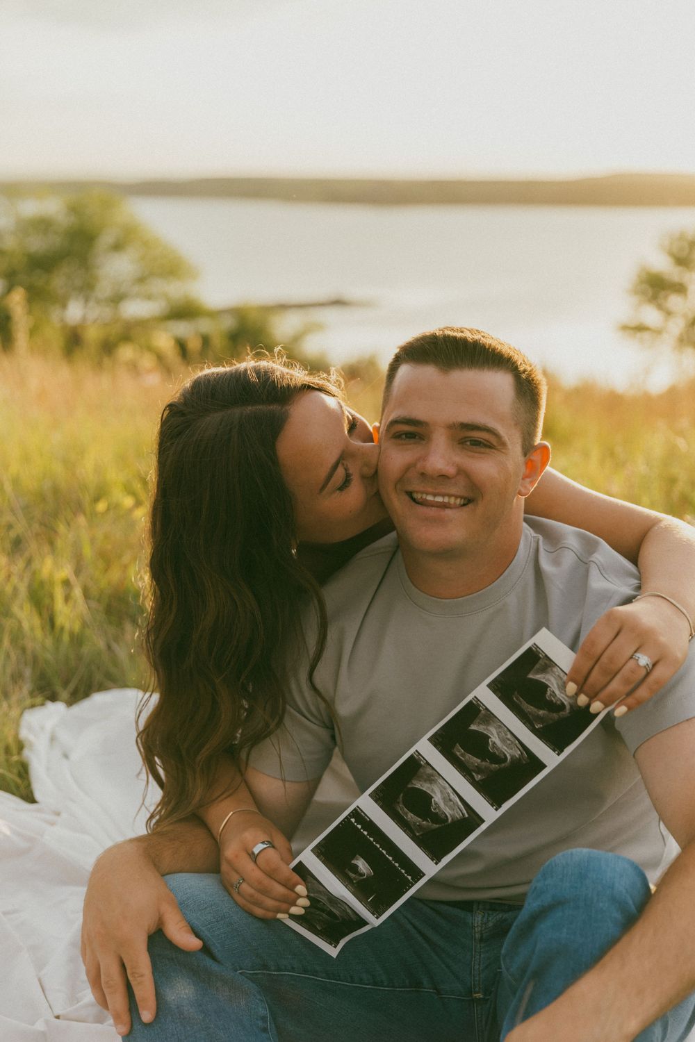 A couple sits together outdoors sharing ultrasound photos during a pregnancy announcement photoshoot at sunset.