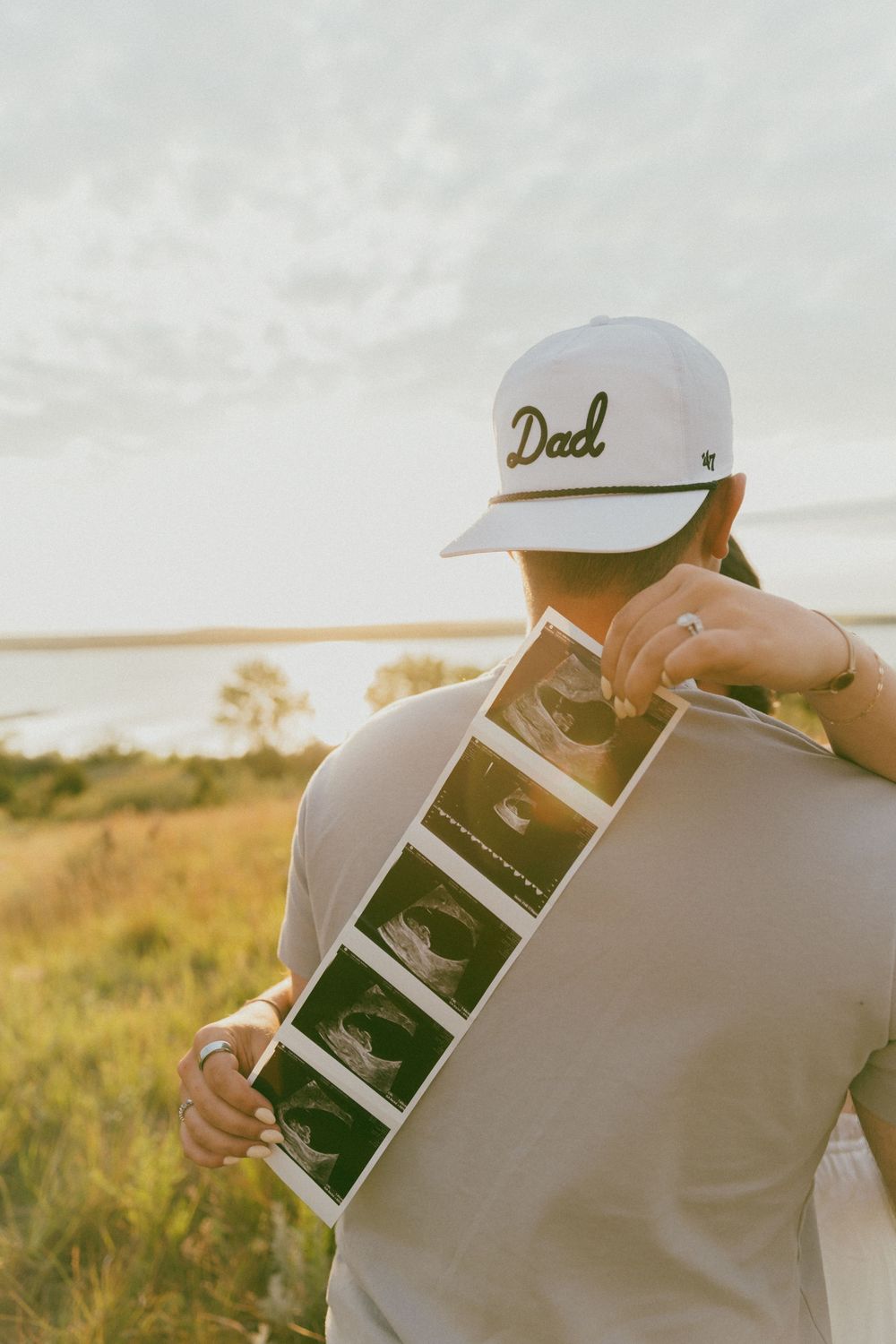 Someone wearing a 'Dad' hat holds up ultrasound images in a sunlit field during golden hour.