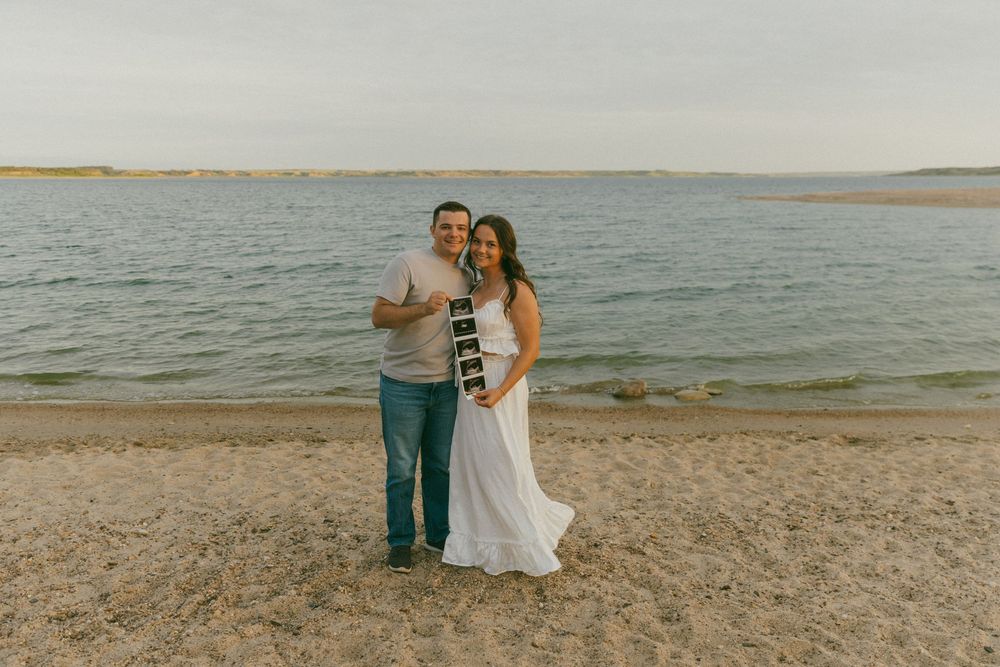 A couple poses together on a sandy beach with a serene lake and distant shoreline in the background.