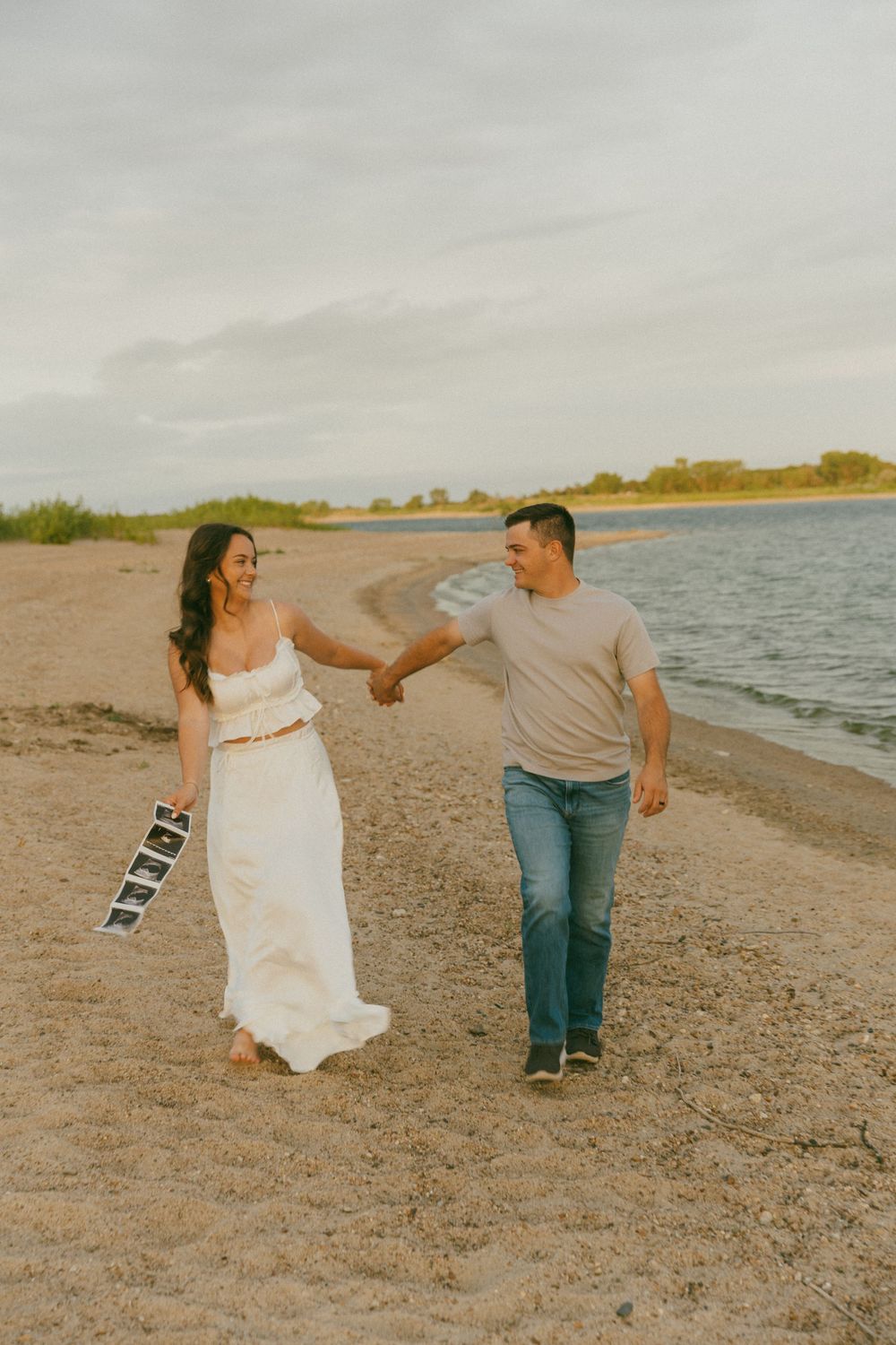 A couple dances playfully on a beach shoreline at sunset, with the woman wearing a flowing white dress.