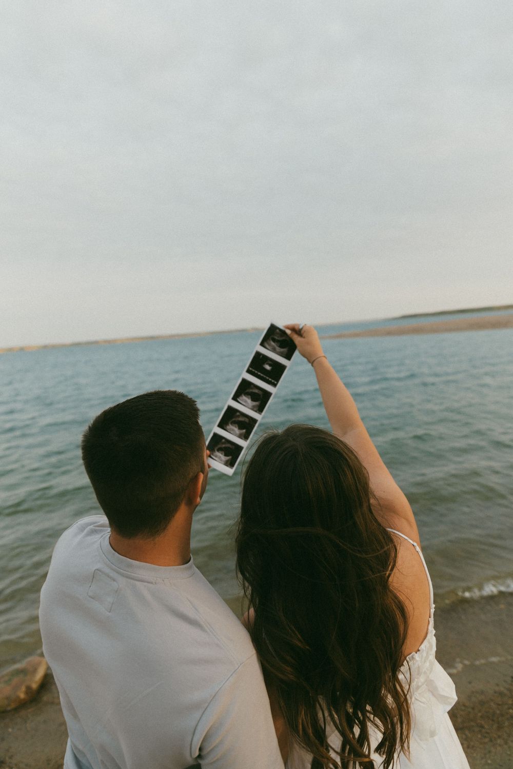A couple sits together on a beach watching the ocean waves while holding ultrasound photos.