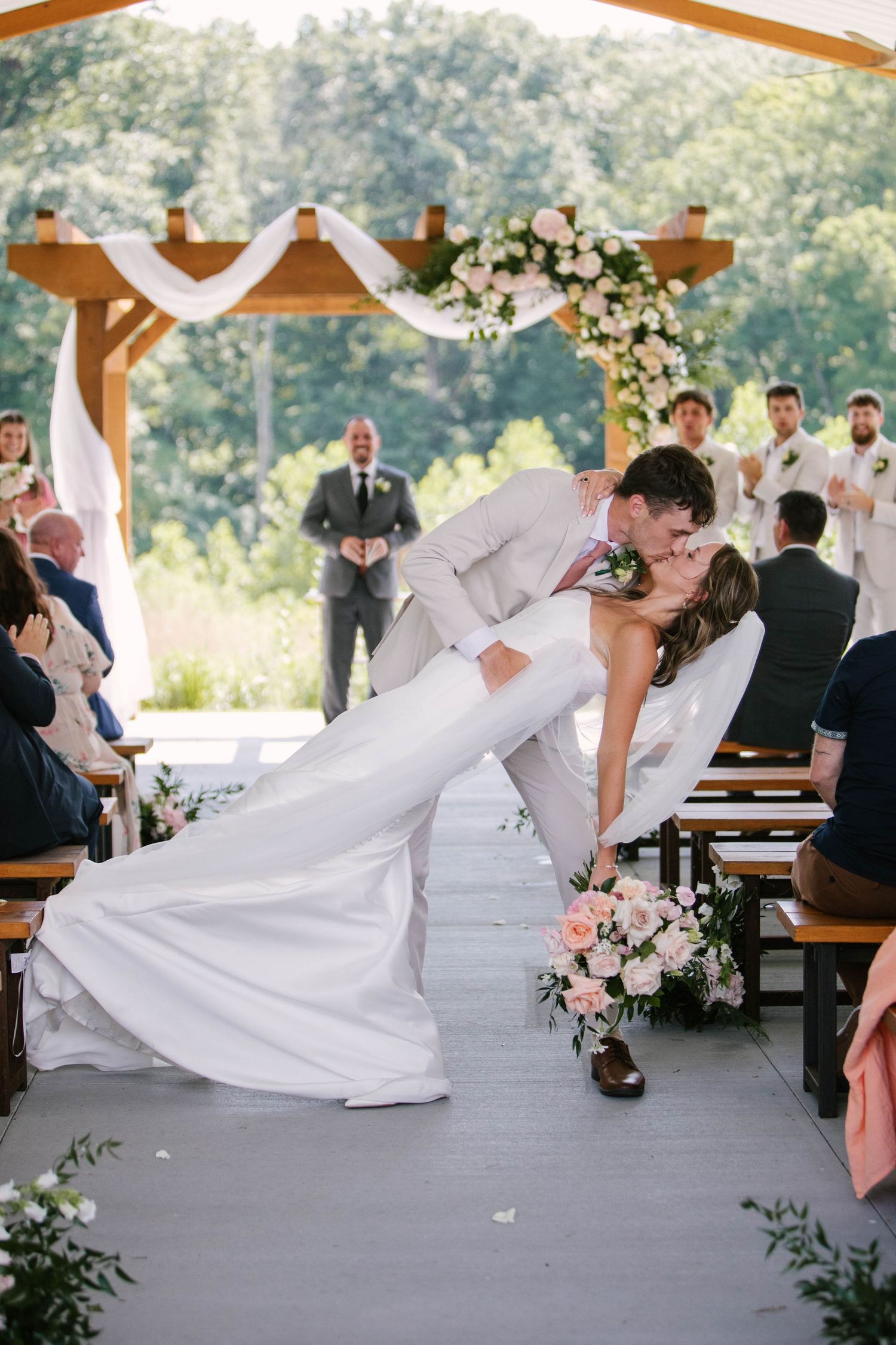 Romantic wedding couple performs dip kiss during first dance under wooden pavilion decorated with white flowers.