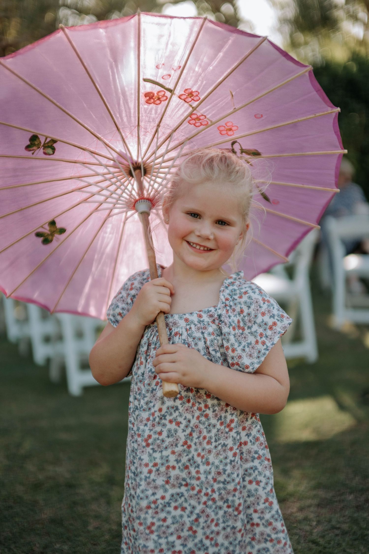 A pink decorative parasol is held outdoors on a sunny day, with a floral dress visible in the scene.